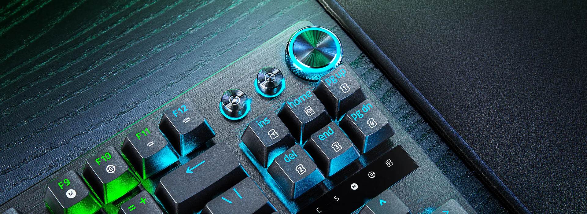 Close-up of a mechanical keyboard with black keys, blue and green backlit labels, and a black mouse pad to the right.
