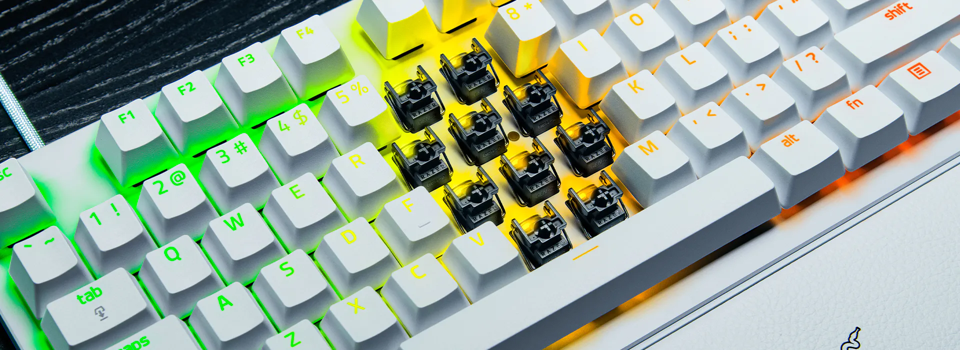 Keyboard with missing keycaps, revealing switches underneath, surrounded by RGB lighting in green, yellow, and orange hues.