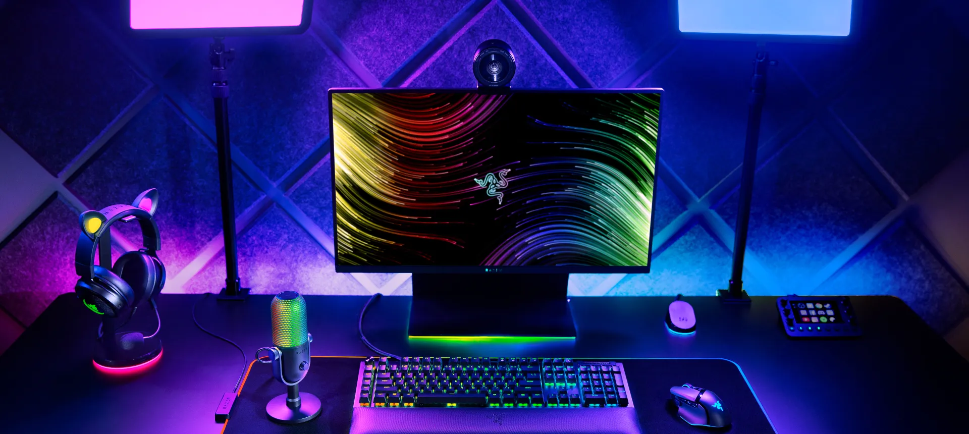 A colorful gaming setup with a Razer monitor, RGB keyboard, mouse, headphones, microphone, and sound mixer, illuminated by vibrant LED lights.