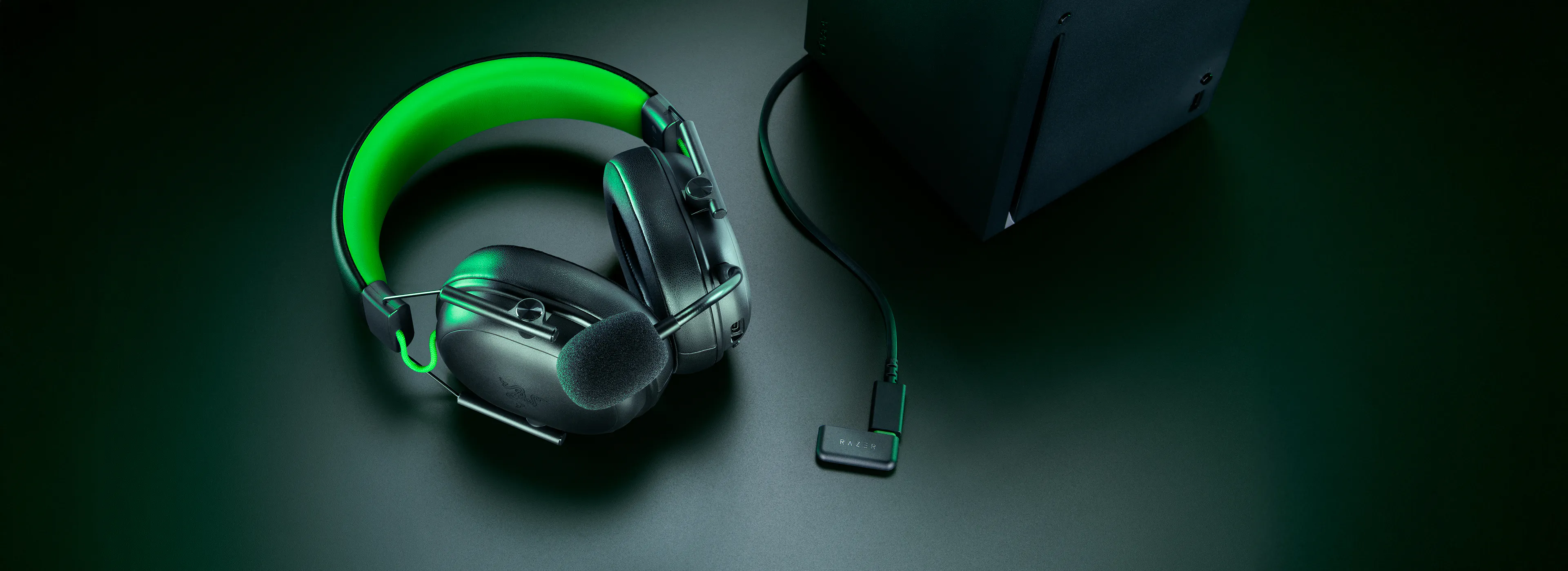 Black gaming headset with a 麦克风, illuminated green accents, connected to a black Razer device on a dark surface.