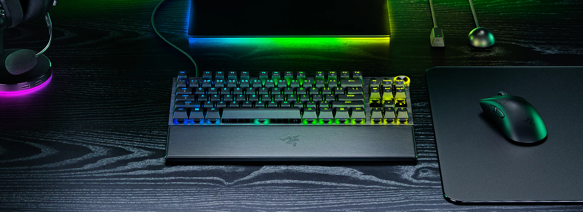 Gaming setup with a RGB mechanical keyboard, Razer mouse on a mat, microphone, headset, and a monitor with rainbow lighting on a black desk.