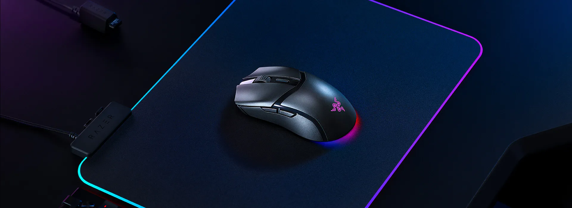 A Razer gaming mouse on a black mouse pad with RGB lighting, connected via a cable, with a wired keyboard partly visible underneath.