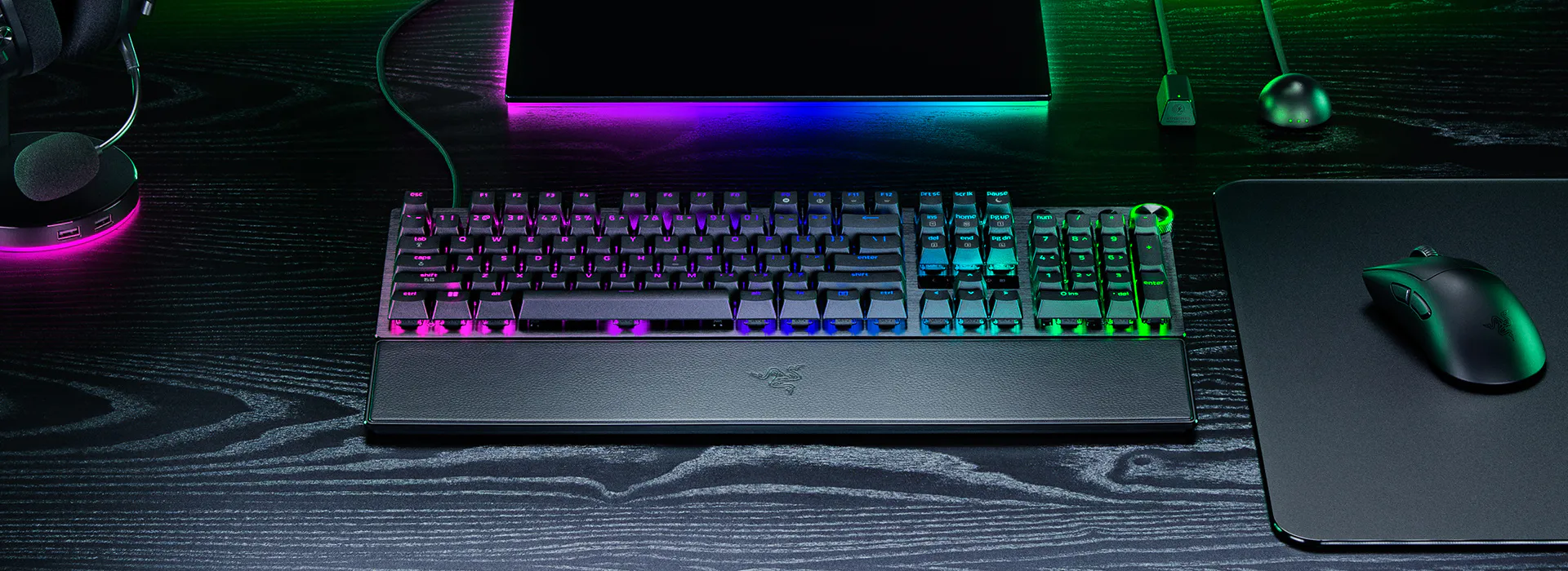 RGB backlit mechanical keyboard, mouse, mouse pad, headset, and monitor on a black wooden desk with neon lighting.