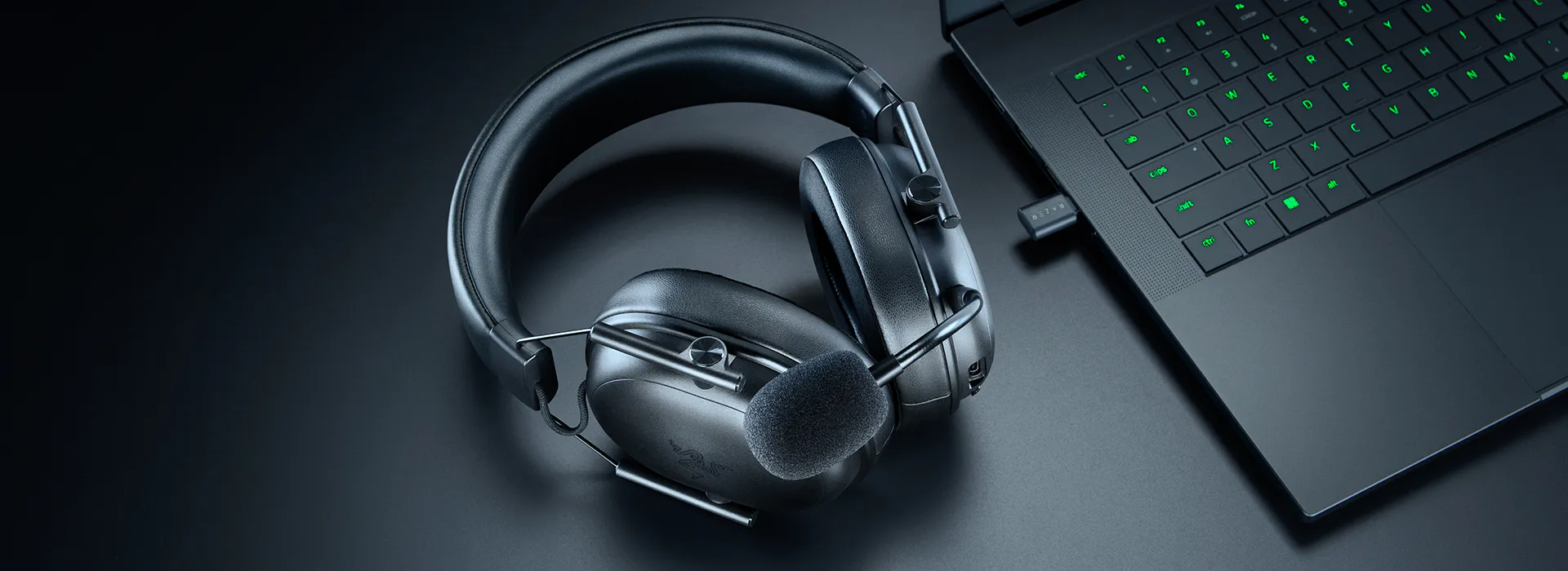 Wireless headset with 麦克风 and a black laptop with green-backlit keyboard on a dark surface.
