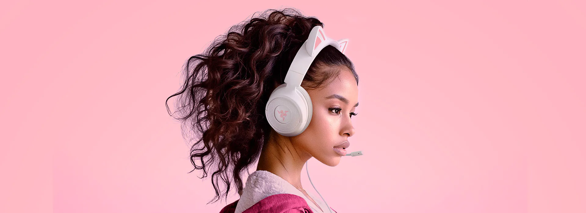 Young woman with curly hair wearing pink and 白色 bunny-eared headphones with a microphone, against pink background.