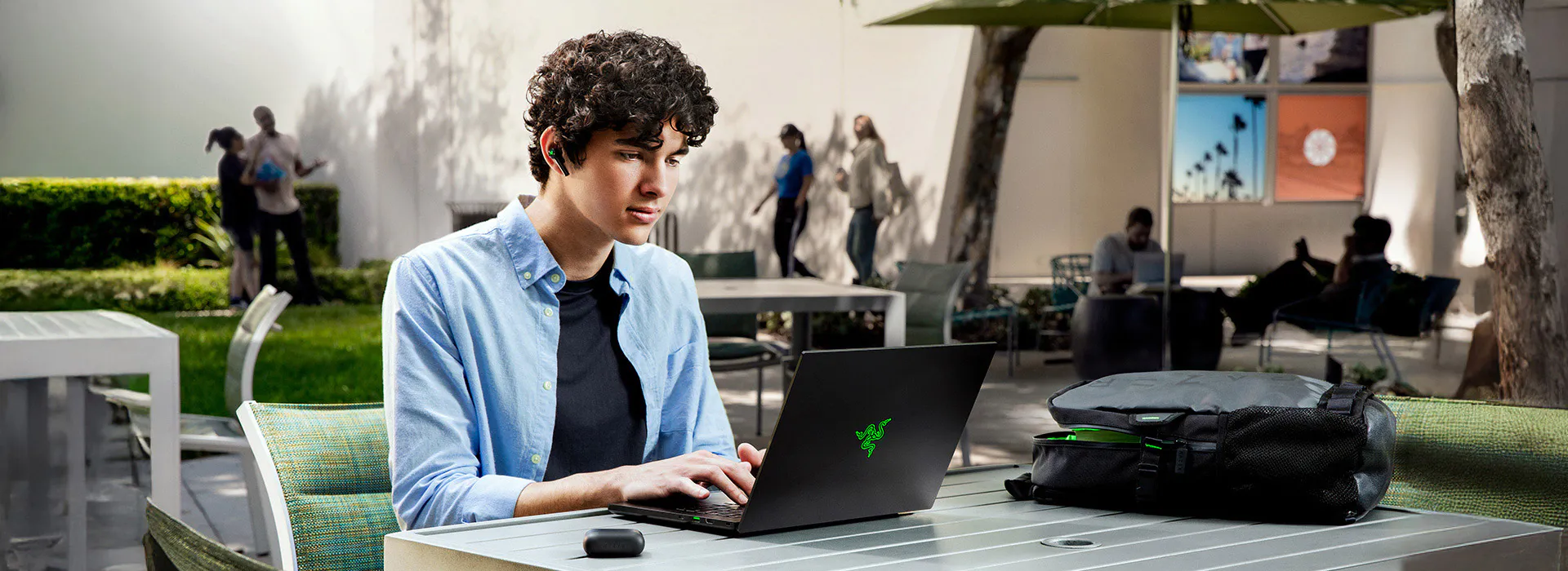 Young man with curly hair and earbuds working on a laptop outdoors, with a black backpack on the table and people in the background.