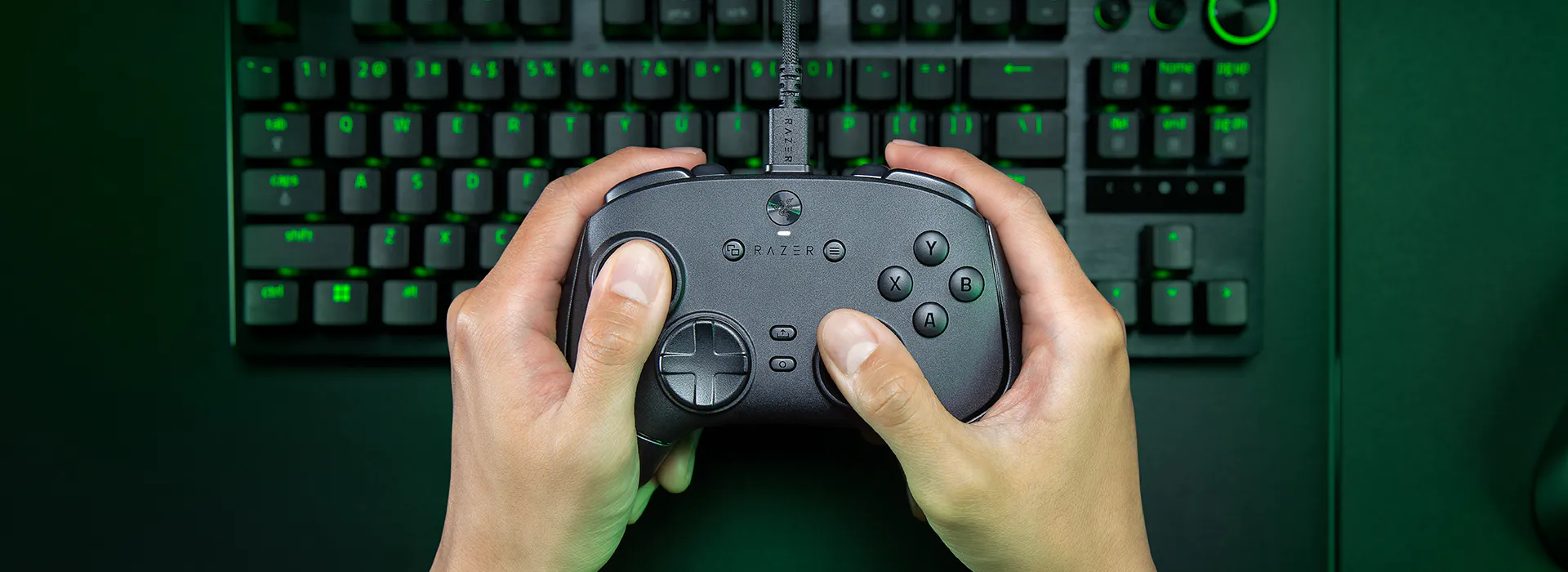 Hands holding a Razer gaming controller above a black gaming keyboard with green backlit keys. The controller has the Razer logo and labels.