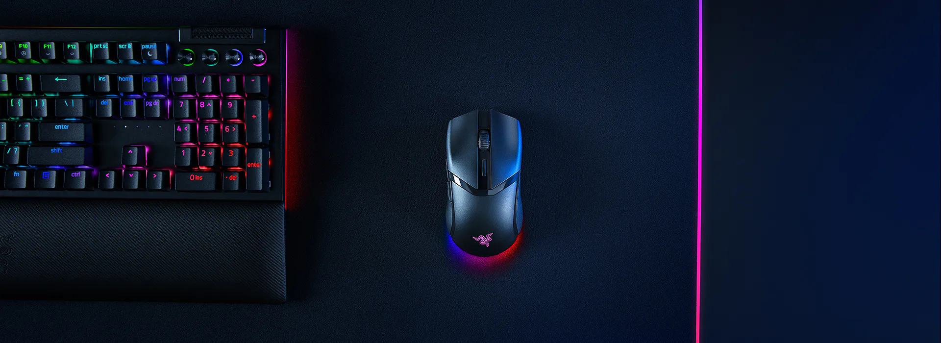 A gaming setup with a black mechanical keyboard with RGB backlighting, a wired mouse with RGB lighting, on a dark gaming mat with pink LED highlights.