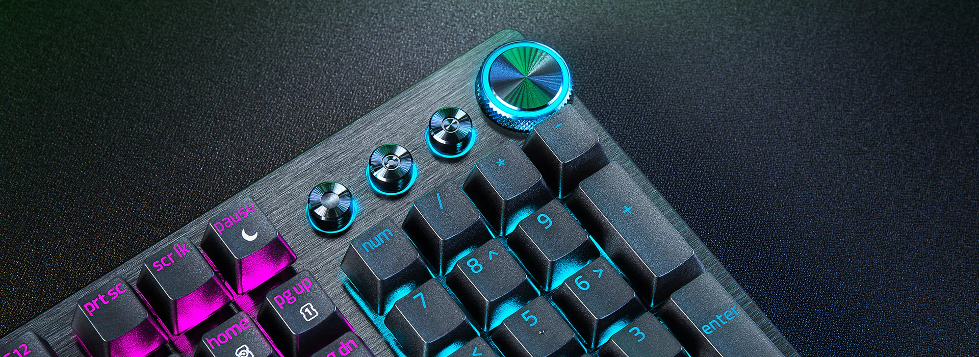 Close-up of a backlit gaming keyboard with colorful keys, metal dials, and textured surface on a dark background.