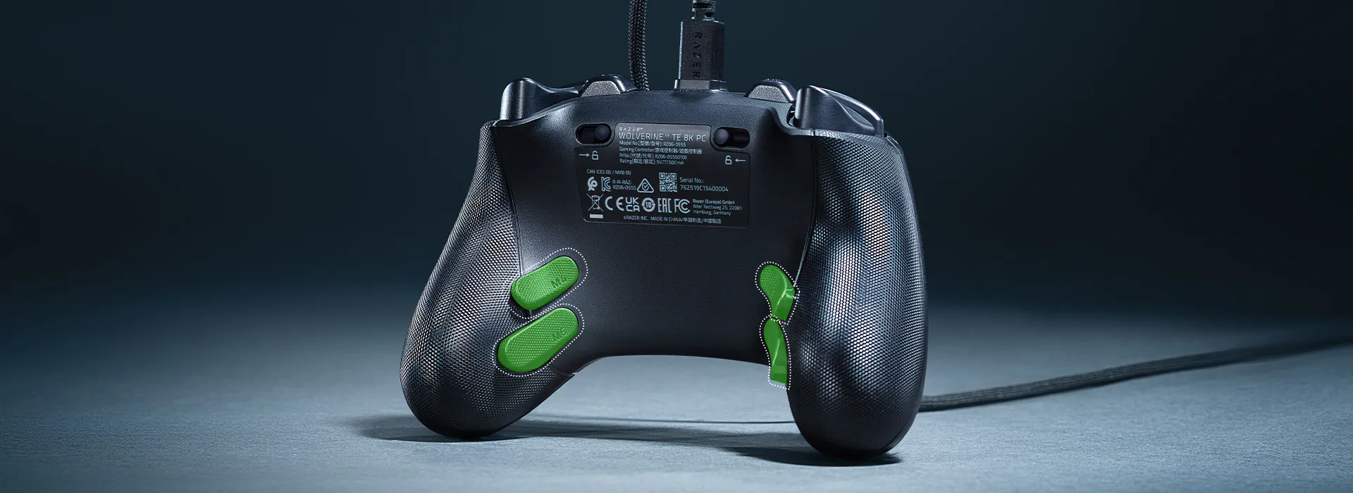 Back of a black Razer Wolverine TE 8K PC controller with green buttons, cable attached, on a gray surface against a dark background.