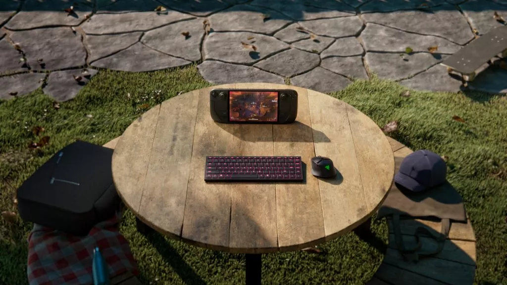 Razer Joro keyboard and Razer Basilisk Mobile mouse arranged with a handheld gaming device on an outdoor round wooden table, surrounded by backpacks and accessories.