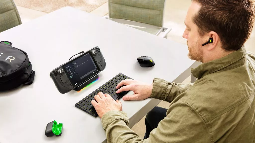 A person using the Razer Joro keyboard paired with a handheld gaming device, with the Razer Basilisk Mobile mouse and earbuds placed on the table nearby.