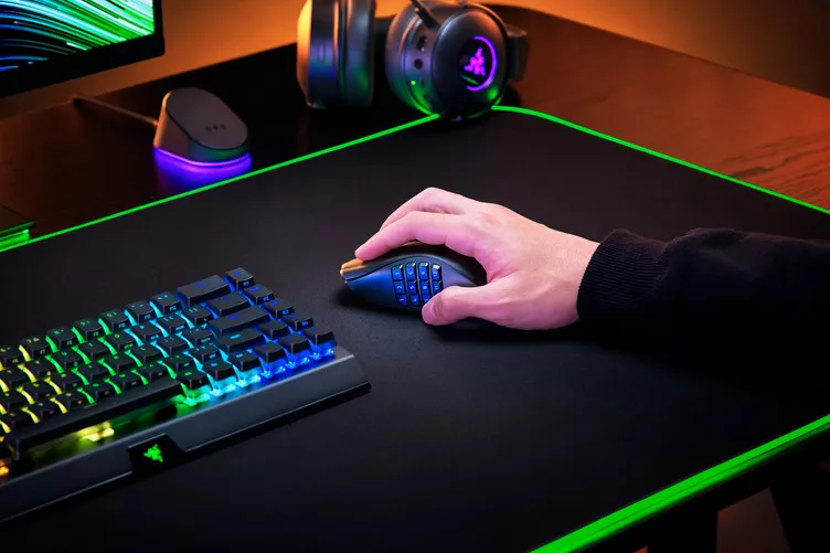 Hand using the Razer Naga V2 Pro on a gaming desk setup with RGB keyboard and headset, highlighting MMO side buttons in use