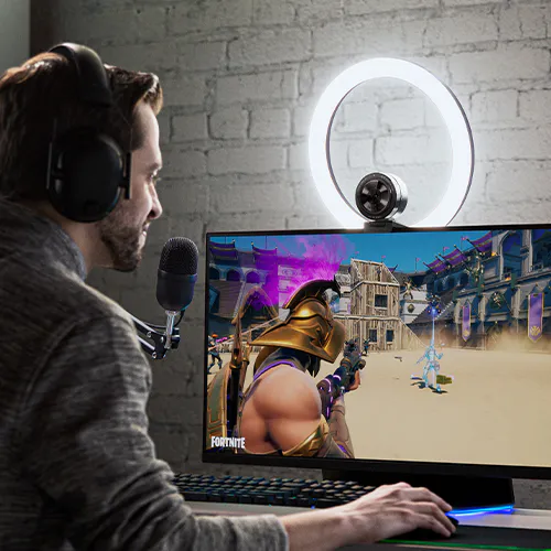 Man with headphones streams Fortnite on a monitor, with a ring light and microphone in a room with a white brick wall.