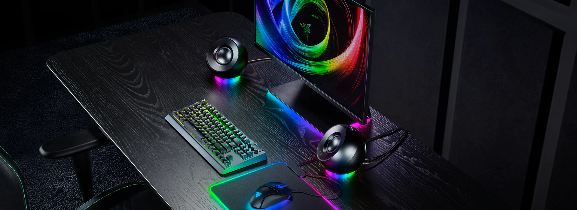 A gaming setup with a colorful RGB monitor, RGB keyboard, mouse, speakers, and RGB-lit mouse pad on a black wooden desk.