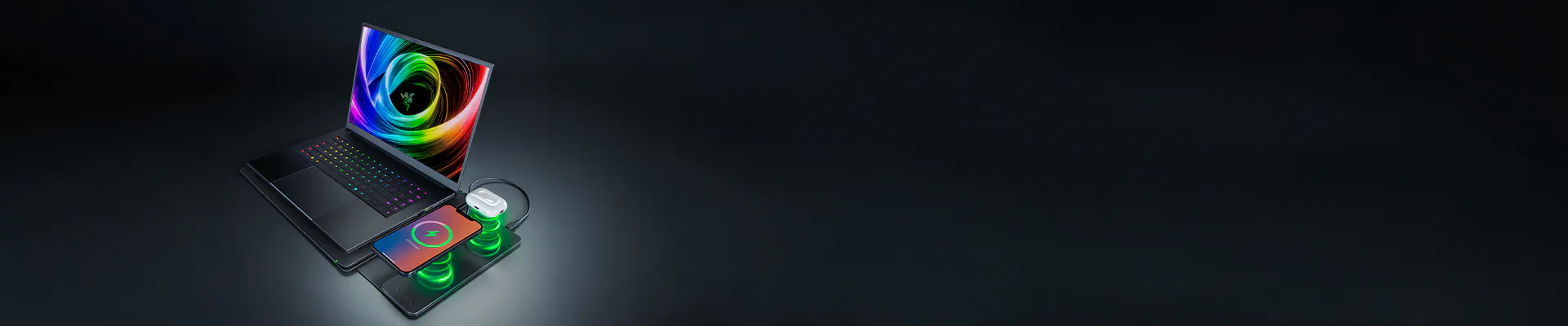 Laptop, phone, and mouse on a wireless charging pad with colorful RGB lighting on a dark surface.