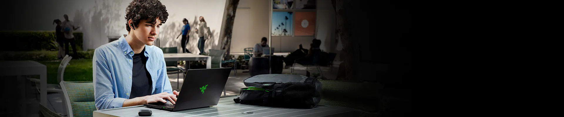 A young person with curly hair working on a black gaming laptop outdoors, with a backpack and a mouse on a table.