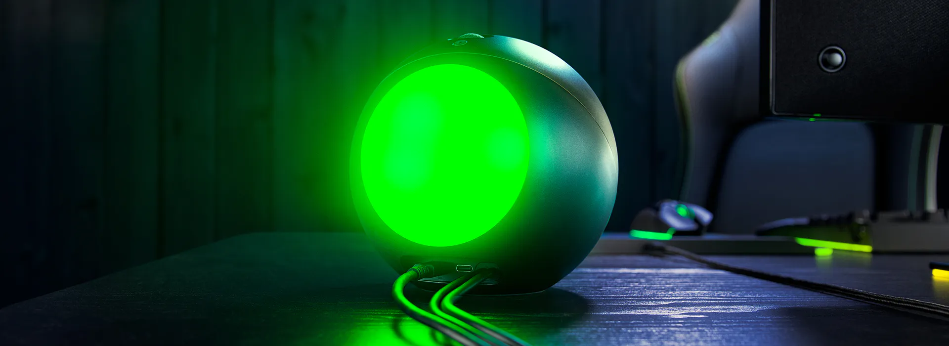 A gaming console with a glowing green light, connected via cables, on a dark desk with a monitor and gaming accessories nearby.