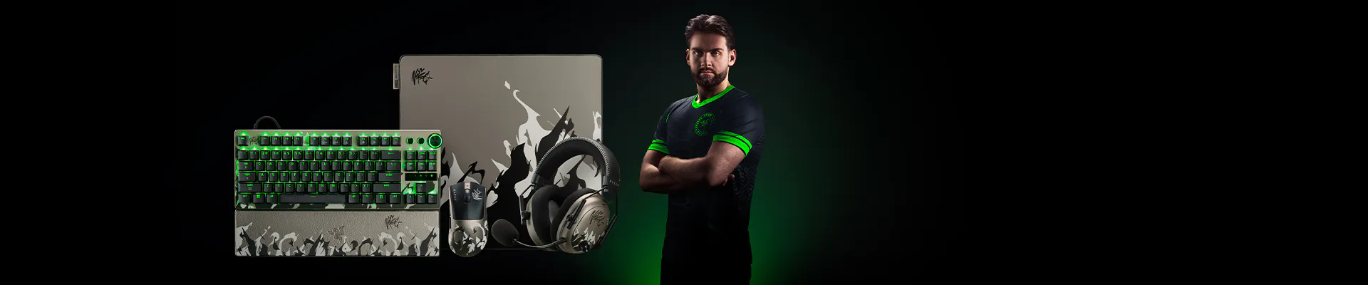 A gamer with a beard stands with arms crossed, surrounded by a gaming keyboard, mouse, headset, and a gaming laptop with tribal art and signatures.