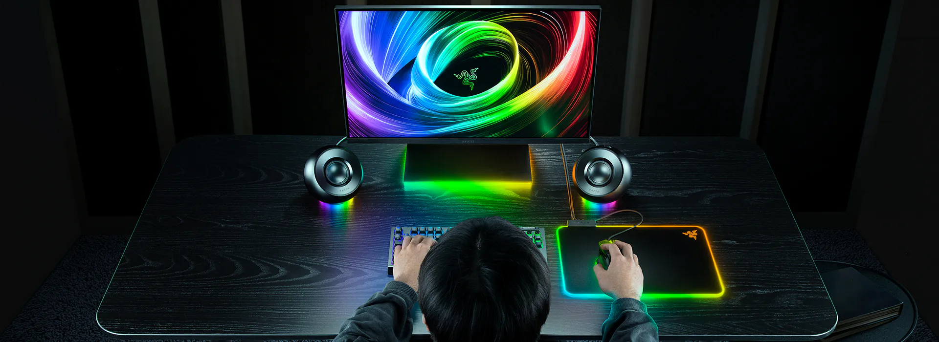 A person using an RGB gaming setup with a monitor, colorful keyb, mouse, illuminated mousepad, speakers, and a dark desk.
