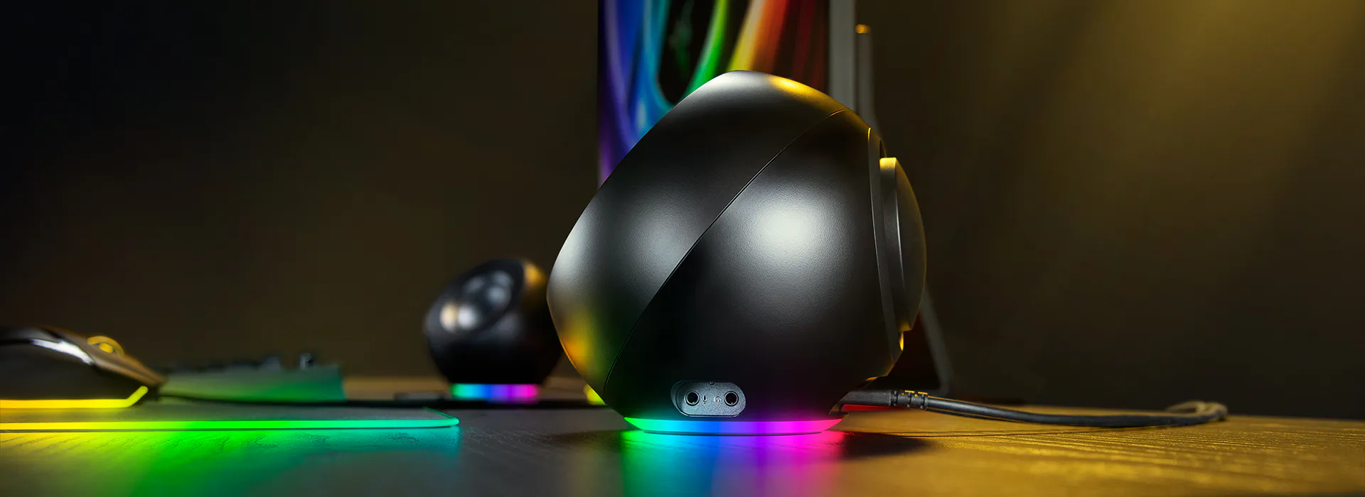 A black gaming mouse, headset, and RGB-lit keyboard on a desk with vibrant LED lighting, arranged against a dark background.
