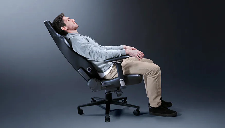 A man relaxes in a black ergonomic office chair, leaning back with eyes closed, against a plain gray background.