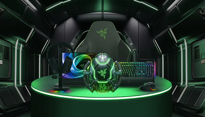 Razer gaming setup: green-lit monitor, a helmet, keyboard, mouse, and Razer logo on the backdrop, all on a round green-lit table in a high-tech room.