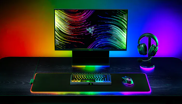 Gaming setup with a Razer monitor displaying colorful lines, a rainbow-backlit keyboard, mouse, large RGB-illuminated mouse pad, and headphones on a black desk.