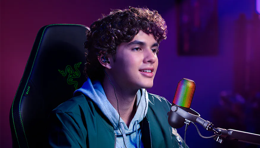 Young man with curly hair smiling at a colorful rainbow microphone, wearing headphones, in a vibrant, dimly lit studio environment.