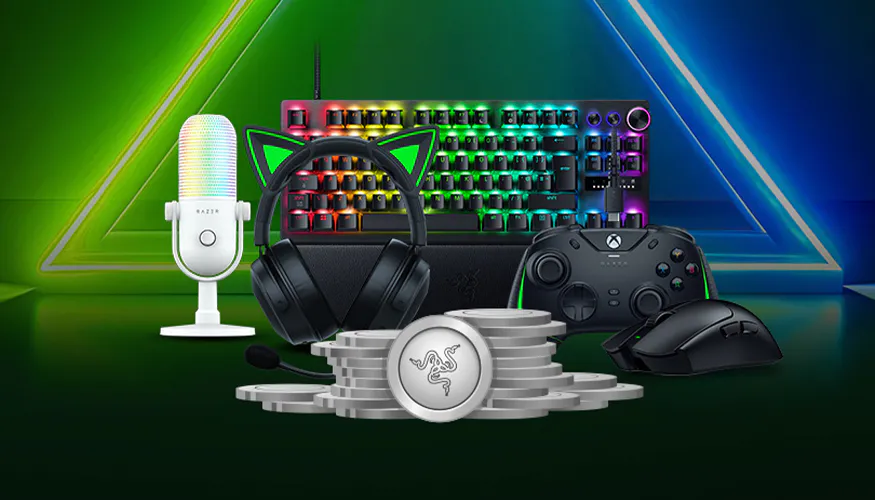 Gaming setup with rainbow-backlit keyboard, Razer microphone, black gaming headset, Xbox controller, Razer mouse, and Razer coins.