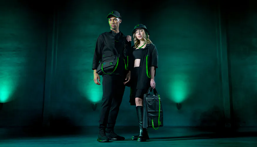 Two models stand in front of a dark green background, dressed in black with neon green accents, holding bags and wearing caps.