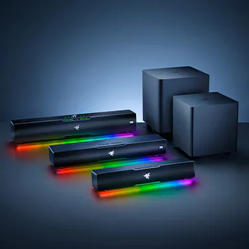 Image shows Razer gaming soundbars with RGB lighting, placed in front of two large black subwoofers on a dark background.
