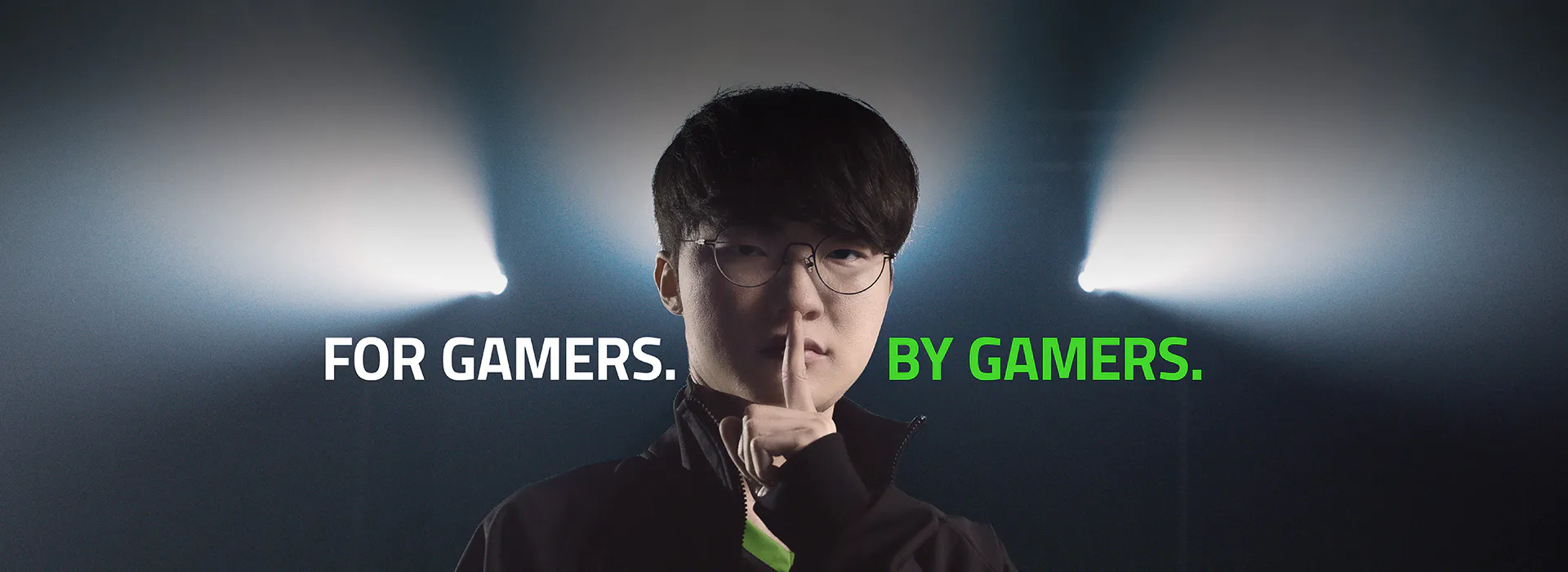 Young man with glasses holds finger to lips, dark background with spotlight, text: "FOR GAMERS. BY GAMERS."