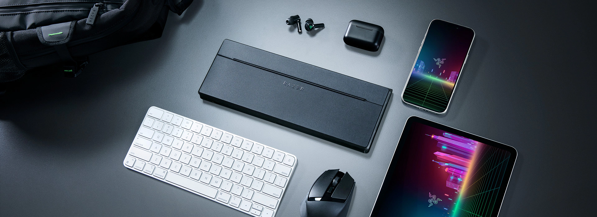 Closed keyboard travel case surrounded by a phone, tablet, keyboard, mouse, earbuds, and backpack on a dark surface.