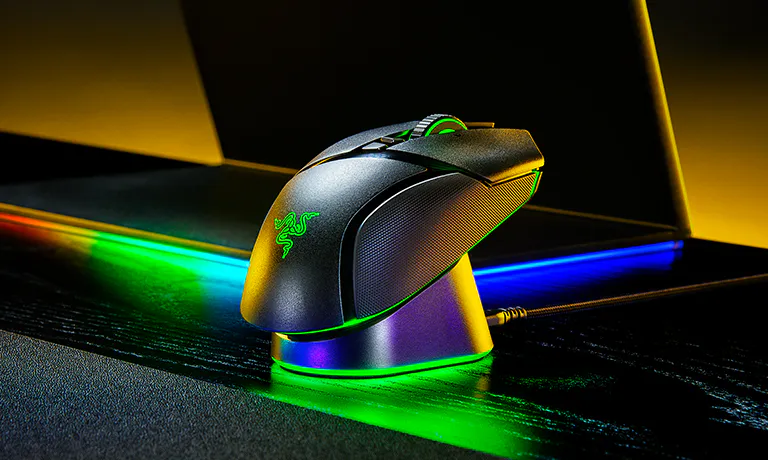 RGB gaming mouse on a desk with colorful lighting, next to a mechanical keyboard, with a laptop in the background.