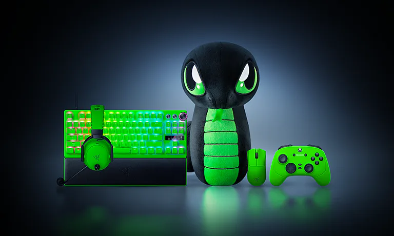 A plush Pokémon toy resembling a snake, surrounded by gaming gear: a green keyboard, headset, mouse, and Xbox controller, all glowing green.