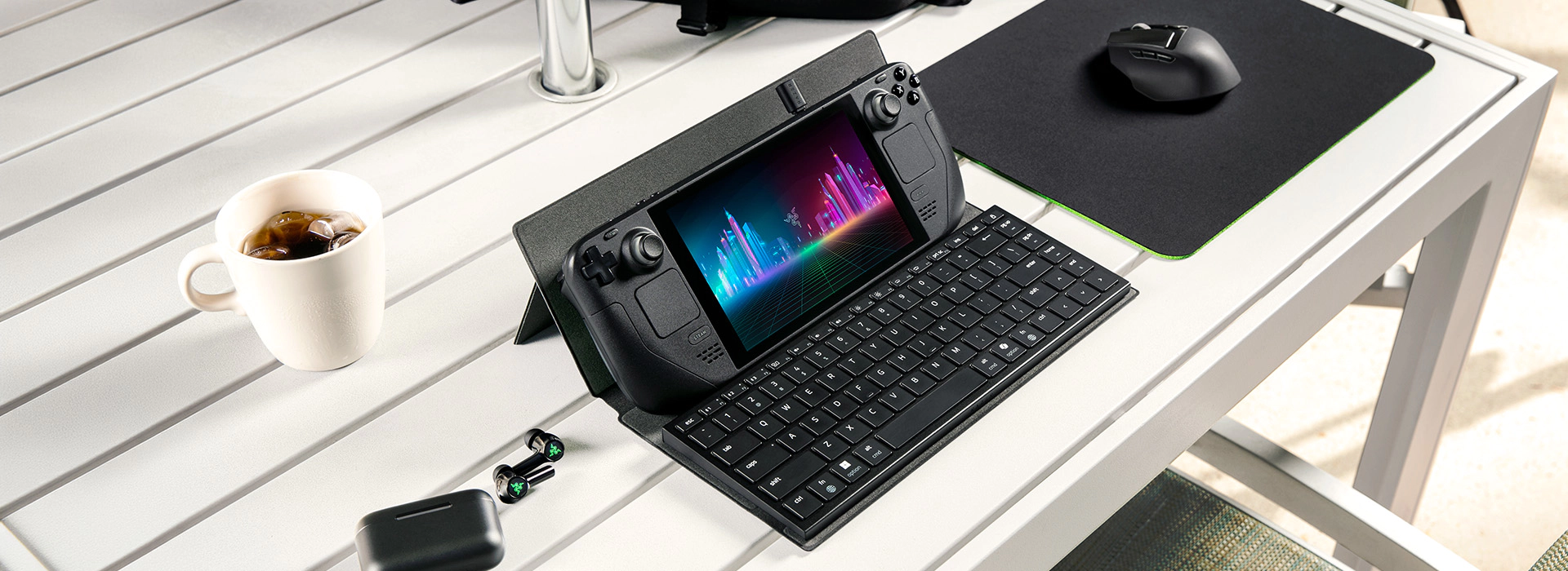 A white outdoor table holds a gaming device with detachable keyboard, a earphone and a black mouse on a mouse pad