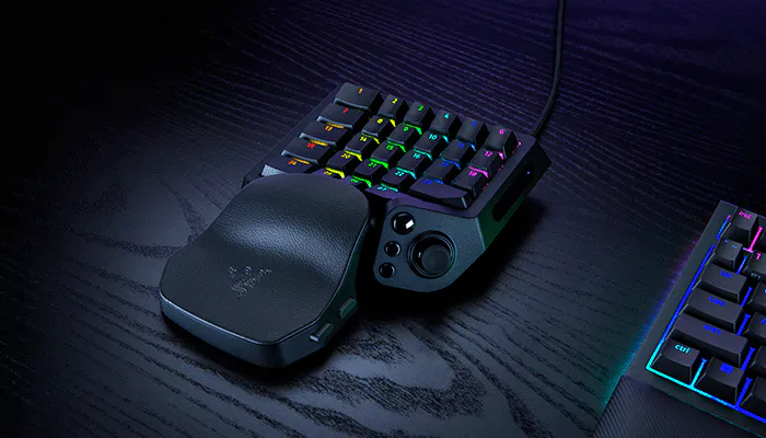 A gaming setup with a black RGB mechanical keyboard, a Razer mouse with a textured grip, and a curved ergonomic keypad on a dark wood desk.