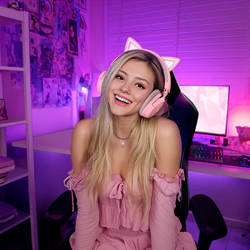 A smiling woman with long blonde hair, wearing pink off-shoulder dress and pink cat-ear headphones, in a neon-lit gaming or streaming room.