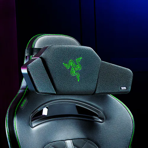 A gaming chair with Razer logo and a headrest pillow, black with green LED accents, in a dark room setting.