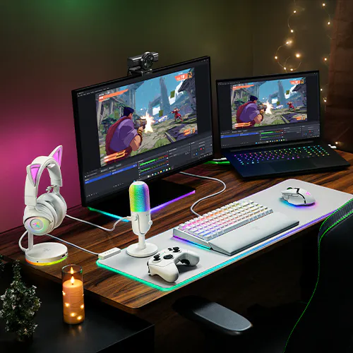 A gaming setup with dual monitors showing a game, RGB keyboard, mouse, game controller, white microphone, cat ear headphones, candle, and decorative lights.