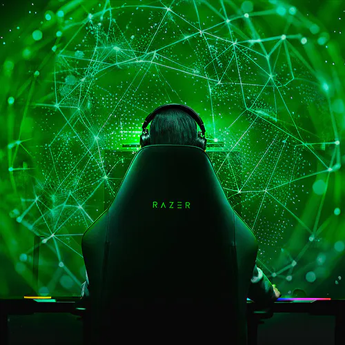 A person with headphones sitting in a Razer gaming chair, facing a digital green network, with the word "RAZER" glowing on the back.