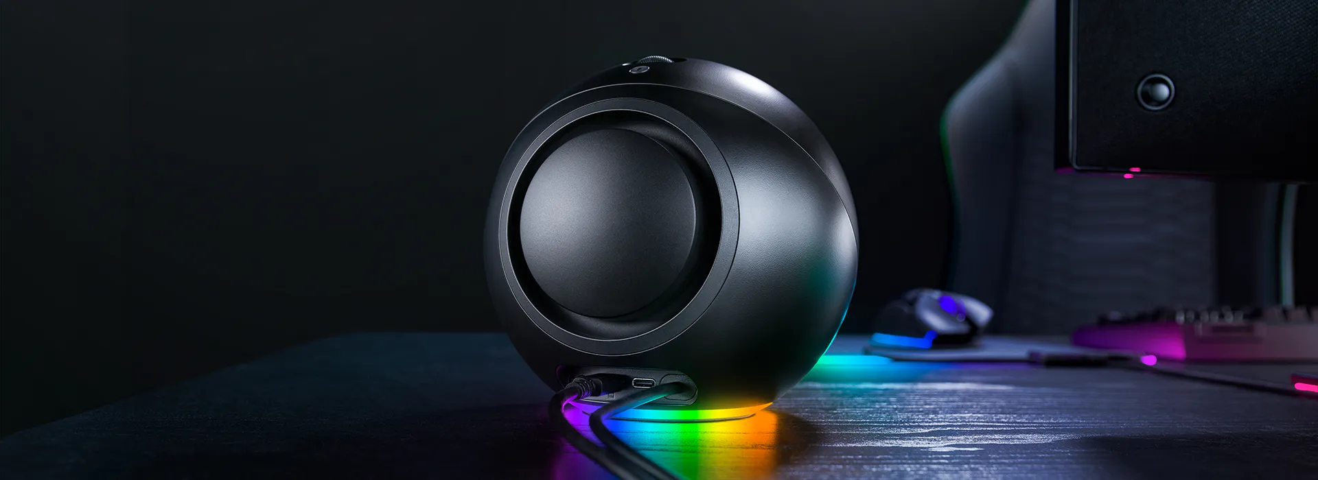 A black spherical speaker with rainbow LED lights at the base, connected to a desk with a monitor and mouse in a dark room.