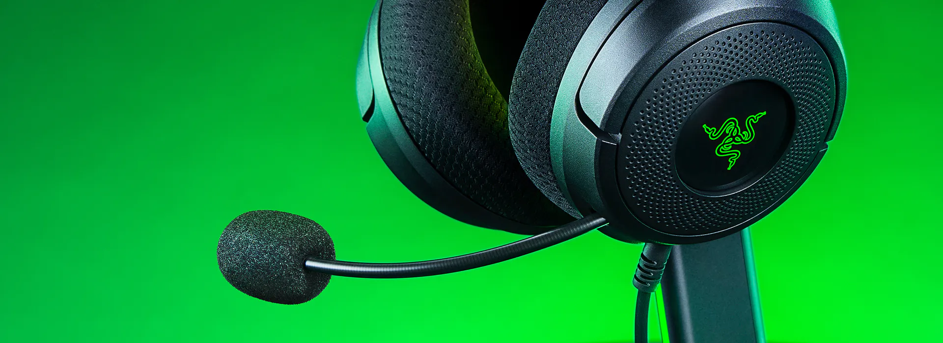 Close-up of Razer gaming headset with mic on black stand against green background, showing the Razer logo on earcup.