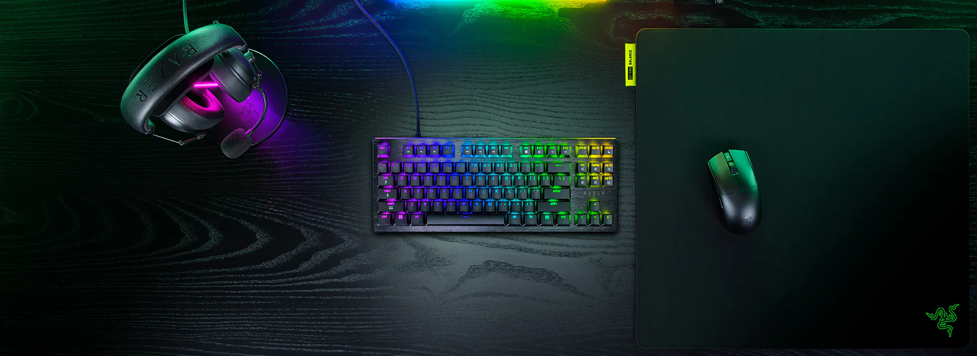RGB backlit keyboard, Razer logo, gaming mouse, headset with Razer logo, mouse pad on a black wooden desk.