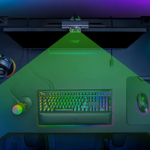 A gaming setup with a keyboard backlit in rainbow colors, a mouse, a pair of headphones, a coffee mug, and a monitor emitting a green laser light.