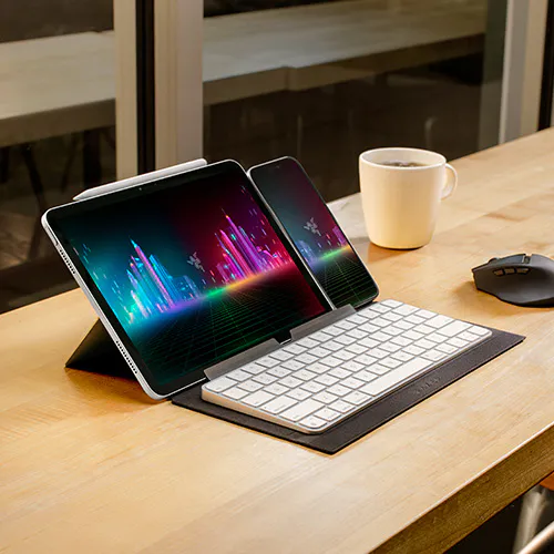A tablet with a keyboard and stylus attached, displaying colorful soundwave graphics, next to a smartphone, coffee mug, and mouse on a wooden desk.