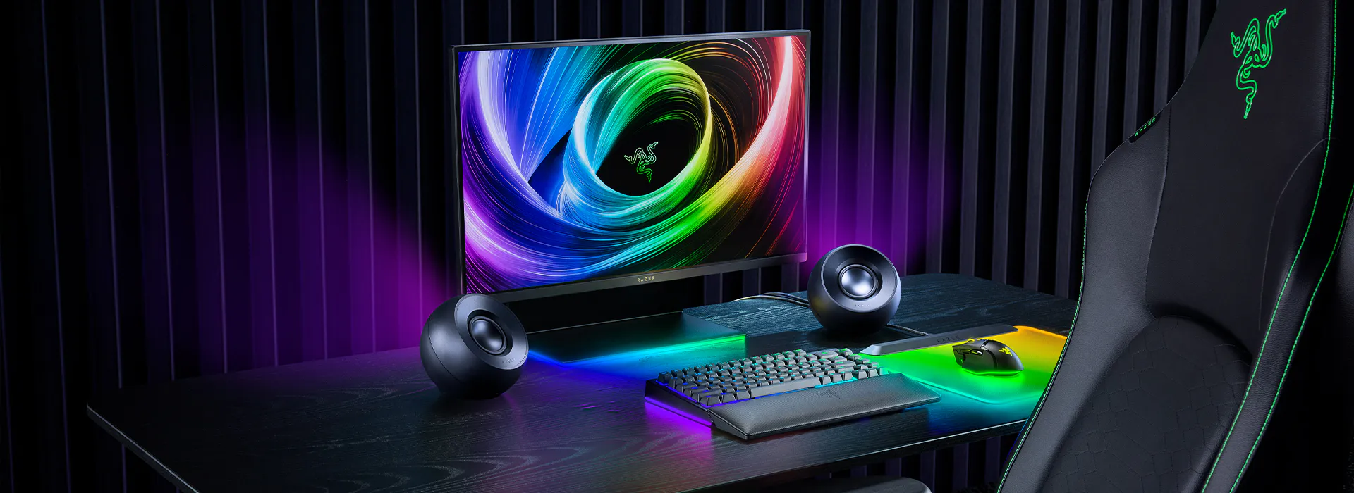 A gaming setup with a Razer monitor, two speakers, a keyboard, mouse, and a gaming chair, all illuminated by RGB lighting in a dark room.