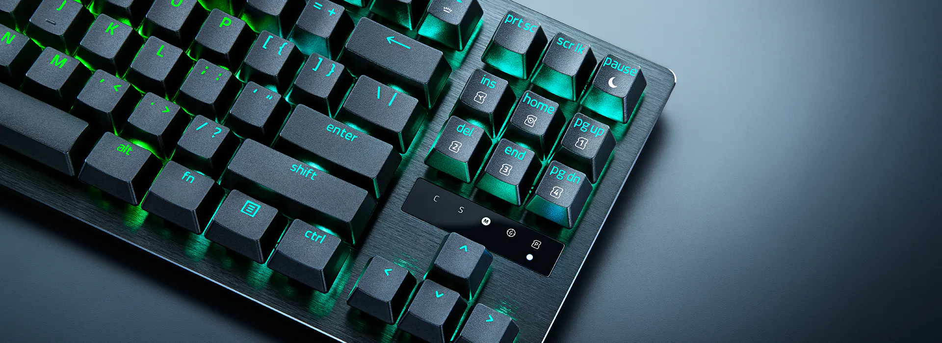 Gaming keyboard with black keys, blue and green backlit labels, and metallic finish on a dark surface.