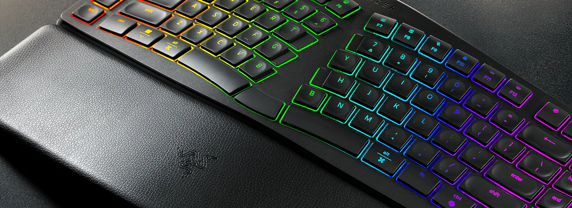 Close-up of a gaming keyboard with RGB backlit keys, positioned above a textured leather wrist rest embossed with the Razer logo.